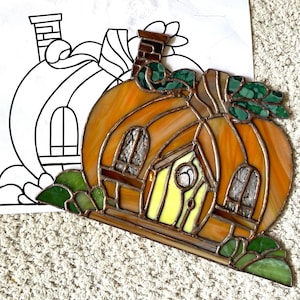 May include: A stained glass pumpkin house with a yellow door, brown windows, and green leaves. The house is made of orange, brown, and green glass.