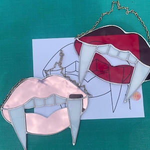 May include: Two stained glass vampire fangs, one red and one pink, with white fangs. The fangs are hanging from a gold chain.