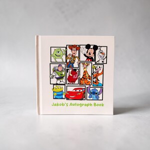 May include: A cream-colored autograph book featuring a grid of cartoon characters, including Mike Wazowski, Woody, Mickey Mouse, and Stitch. The book is labeled "Jakob's Autograph Book" in green text.