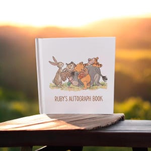May include: A white autograph book with a colorful illustration of cartoon characters, including a rabbit, owl, bear, and tiger. The book is labeled "RUBY'S AUTOGRAPH BOOK" in brown text. The book is on a wooden surface with a blurred background.