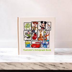 May include: A white autograph book featuring a collage of Disney characters, including Mickey Mouse, Buzz Lightyear, and Lightning McQueen. The book's cover has the text "Thatcher's Autograph Book" in green. The book is on a white marble surface.