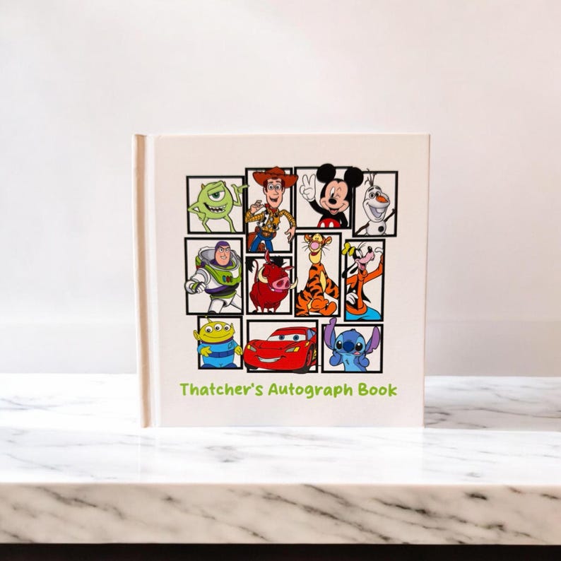 May include: A white autograph book featuring a collage of Disney characters, including Mickey Mouse, Buzz Lightyear, and Lightning McQueen. The book's cover has the text "Thatcher's Autograph Book" in green. The book is on a white marble surface.