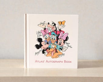 May include: A white hardcover autograph book featuring a colorful illustration of Disney characters including Mickey Mouse, Minnie Mouse, Donald Duck, and Goofy. The title "Atlas' Autograph Book" is printed in red at the bottom.