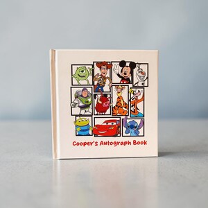 May include: A cream-colored autograph book featuring a collage of Disney characters, including Mickey Mouse, Buzz Lightyear, and Lightning McQueen. The title "Cooper's Autograph Book" is printed in red script.