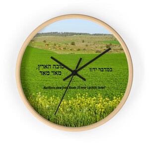 May include: A wooden wall clock with a green field and yellow flowers in the background. The clock face features Hebrew text that reads "במדבר יד:ז טובה הארץ, מאד מאד" and "Northern view from Route 35 near Lachish, Israel".
