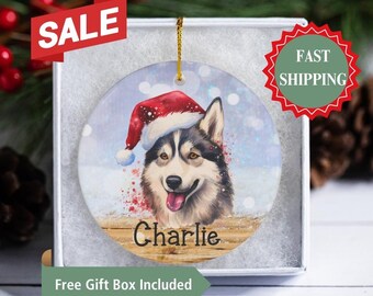 May include: A white ceramic Christmas ornament with a painting of a Siberian Husky wearing a red Santa hat. The ornament has a snowy background and the name "Charlie" is written on a wooden sign at the bottom.