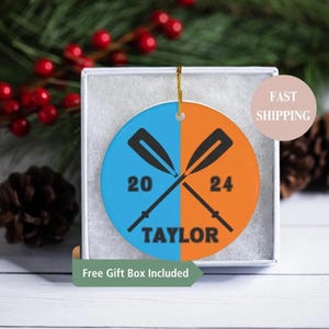 May include: A round, blue and orange ceramic ornament with two crossed oars, the numbers 20 and 24, and the name "TAYLOR".