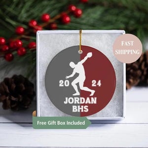 May include: A round, two-toned ornament with a gray and maroon color scheme. A white silhouette of a discus thrower is featured, along with the numbers "20" and "24", the name "JORDAN", and the initials "BHS". The ornament is in a gift box.