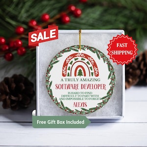 May include: A white ceramic Christmas ornament with a green wreath and a rainbow design. The ornament reads "A truly amazing software developer is hard to find, difficult to part with and impossible to forget." The name "Alexis" is printed below the text.