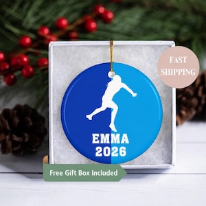 May include: A round ceramic ornament featuring a split blue and light blue background with a white silhouette of a person running. The text EMMA and 2026 are printed in white below the figure. A gold string is attached to the top for hanging.