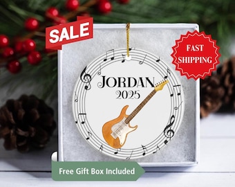 May include: A round, white ceramic ornament featuring a watercolor illustration of an electric guitar. The ornament has musical notes around the edge, with the name "JORDAN" and the year "2025" printed in black. The ornament is in a gift box.
