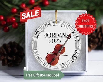 May include: A white round ornament with a violin illustration, musical notes, and the name "JORDAN" and the year "2025". The ornament is in a gift box. A red "SALE" tag and a "FAST SHIPPING" tag are visible.