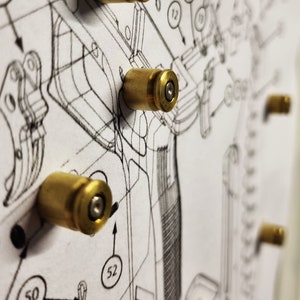 Puede incluir: Primer plano de un diagrama con varias vainas de bala de latón adjuntas. El diagrama parece ser un dibujo técnico de un arma de fuego, con varios componentes numerados y líneas. Las vainas están espaciadas uniformemente.