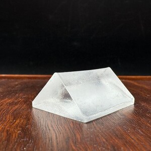 Può includere: Un prisma di vetro trasparente con forma triangolare, posto su una superficie di legno.