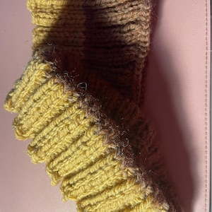 Puede incluir: Bufanda tejida a mano, de color amarillo mostaza y marrón. La bufanda está hecha de hilo grueso y tiene una textura acanalada. Los bordes son festoneados, lo que añade un toque decorativo. La bufanda está doblada, mostrando los diferentes colores y texturas.
