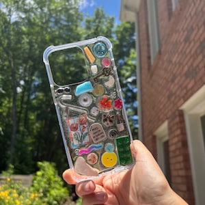 May include: A clear phone case with a variety of colourful objects embedded inside, including a peace sign, a key, a flower, a button, a small cat figurine, and a piece of paper with the text "MRS. ANITA HOWARBY 1901-1981".