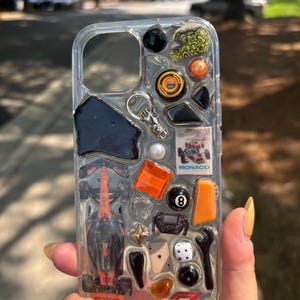 以下が含まれることがあります： 埋め込み装飾要素を備えた透明な携帯電話ケース。 ミニチュアのレーシングカー、オレンジと黒のアクセント、黒の8ボール、マクラーレンのロゴが含まれています。 ケースは手で持たれています。