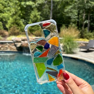 May include: A clear phone case with a mosaic design made of colorful glass pieces. The case is made of a clear material and the glass pieces are embedded in the case. The glass pieces are in various shapes and sizes and are arranged in a random pattern. The colors of the glass pieces are red, orange, yellow, green, blue, purple, and brown.