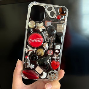 May include: A clear phone case with a variety of colorful and metallic embellishments, including a Coca-Cola logo, a red heart, a star, a dice, and a wrench.