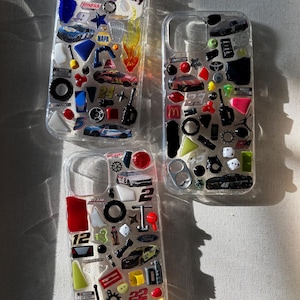 Puede incluir: Tres fundas transparentes para teléfono decoradas con miniaturas de coches de carreras y elementos temáticos de la NASCAR. Cada funda presenta una variedad de elementos coloridos, incluyendo modelos de coches, logotipos y otros accesorios relacionados con las carreras. Las fundas están dispuestas sobre una superficie neutra.