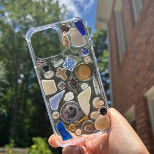 May include: A clear phone case with a variety of small objects embedded inside, including shells, keys, a small plate, a cross, a dice, and a blue eye charm.