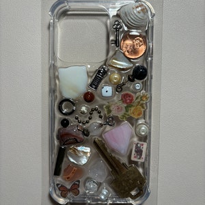 May include: A clear phone case with a variety of small objects embedded inside, including a key, a seashell, a coin, a butterfly, a dice, and a playing card. The objects are arranged in a random pattern and are visible through the clear case.