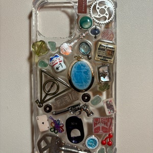 Könnte beinhalten: Eine transparente Handyhülle mit verschiedenen Charms und Schmuckstücken, die darin eingebettet sind. Die Charms umfassen eine kleine Katzenfigur, ein kleines Banjo, einen Schlüssel, einen Flaschenöffner, eine Spielkarte, einen kleinen Kompass, eine kleine Pistole, ein kleines Stück Papier mit dem Text "THORNTON 17", ein kleines Stück Papier mit dem Text "The Daily Precept EXCLUSIVE", ein kleines Stück Papier mit dem Text "MASS BREAKOUT AZKARAN", ein kleines Stück Papier mit dem Text "US AIR MAIL" und ein kleines Stück Papier mit dem Text "Nu".