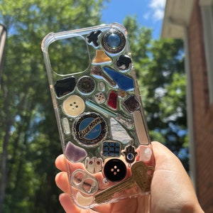 May include: A clear phone case with a variety of small objects embedded in the resin. The objects include buttons, bottle caps, keys, dice, and a small blue moon logo.