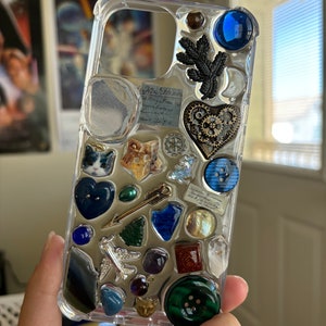 May include: A clear phone case with a variety of colorful and textured embellishments, including a heart, a cat, a star, a leaf, a button, and a plane. The case is decorated with a variety of colors, including blue, green, gold, and silver.