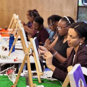 May include: A group of people are painting at easels in a well-lit room. Each person is focused on their canvas, holding a paintbrush. The easels are wooden, and the canvases are white. The scene suggests an art class or workshop.