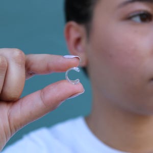 May include: A small, clear, C-shaped plastic ring adjuster designed to fit onto the band of a ring. The transparent, flexible component features a curved design that allows it to wrap around a ring band to provide a snugger fit for the wearer.