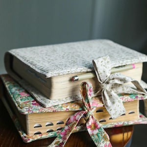May include: Two books with floral patterned covers are stacked on a wooden surface. The top book has a light green and white pattern, while the bottom book has a colorful floral design. A pencil rests on the top book, and both are tied with decorative ribbons.