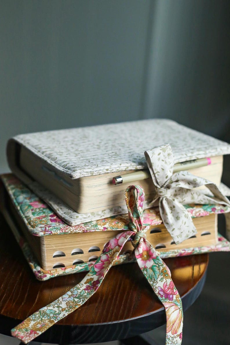 May include: Two books with floral patterned covers are stacked on a wooden surface. The top book has a light green and white pattern, while the bottom book has a colorful floral design. A pencil rests on the top book, and both are tied with decorative ribbons.