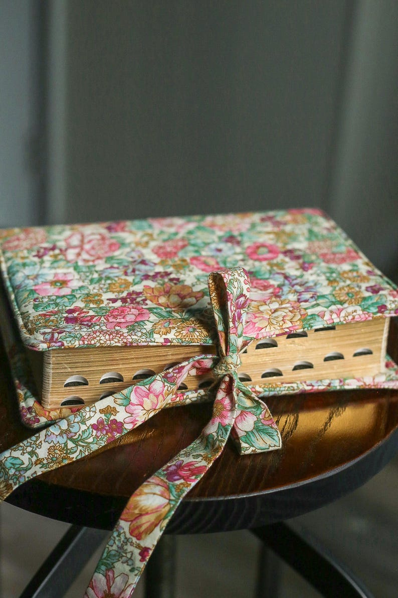 May include: A floral-covered book with a matching ribbon tied around it. The book's cover and ribbon feature a pattern of pink, peach, and green flowers on a light background. The book is resting on a wooden surface.