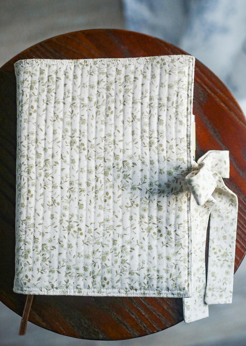 May include: A floral-patterned journal with a white background and olive green flowers. The cover is quilted and tied with a matching ribbon. The journal is resting on a dark wooden surface.