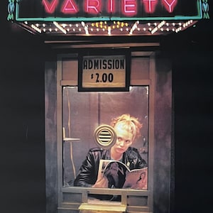 以下が含まれることがあります： VARIETYと書かれたネオンサインがあるチケットブースの映画ポスター。上部にはRonald Chammah présente、下部にはUN FILM DE BETTE GORDONの文字。ブースの中に座った人物が、ADMISSION $2.00と書かれた看板の下で雑誌を読んでいる。