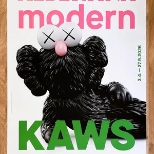 May include: Poster with a black KAWS figure, white eyes with crosses, and a pink nose. The poster reads "ALBERTINA modern" in pink and "KAWS ART & COMIX" in green. The date "3.4. - 27.9.2026" is also displayed.