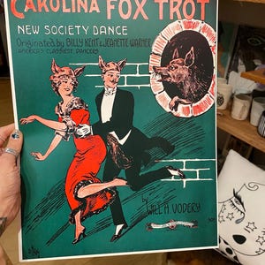 May include: A vintage poster titled "Carolina Fox Trot" with text and illustrations. The poster features a couple dancing, wearing fox-themed headwear, and a fox peering from a circular opening. The color palette includes red, green, and black.