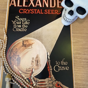 May include: A vintage poster advertising a fortune teller named Alexander, the Crystal Seer. The poster features a skeletal hand holding a crystal ball, with a skull in the top right corner. The text on the poster reads "Alexander Crystal Seer Sees Your Life from the Cradle to the Grave."
