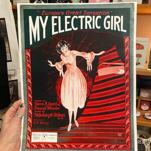 May include: Vintage sheet music cover titled "My Electric Girl." The artwork features a woman in a pink dress with outstretched arms against a black and red background. The text includes the names of the songwriters and publisher.
