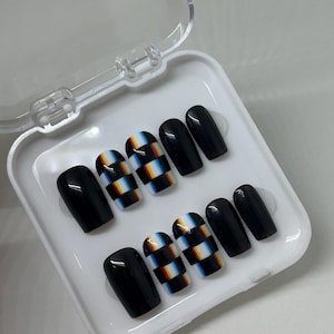 May include: A set of black and patterned press-on nails in a white plastic case. The nails alternate between solid black and a checkered design with red, yellow, blue, and white stripes. The nails are oval-shaped.