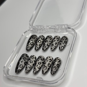 Könnte beinhalten: Ein Set aus zehn silber-schwarzen Leopard-Print-Press-On-Nägeln in einer transparenten Kunststoffbox. Die mandelförmigen Nägel haben eine glitzernde silberne Basis mit schwarzen Flecken. Die Box ist geöffnet und zeigt das komplette Set.