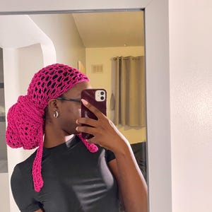 Puede incluir: Un gorro de pelo de ganchillo rosa con una lazada larga y suelta. El gorro lo lleva una persona de piel oscura y pelo corto. La persona lleva una camisa negra.
