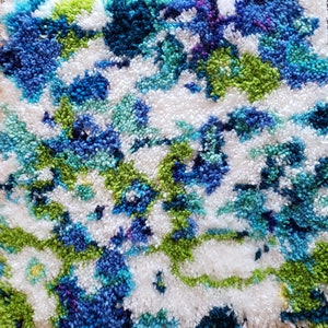 May include: Close-up of a textured rug with a floral pattern. The rug features a mix of white, blue, green, and purple hues. The design is abstract, with the colours blending together to create a soft, inviting look. The rug's texture adds depth and visual interest.