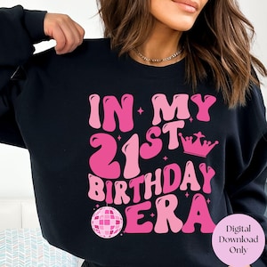 May include: Black sweatshirt with pink text that reads "In My 21st Birthday Era" with a pink disco ball and crown graphic.