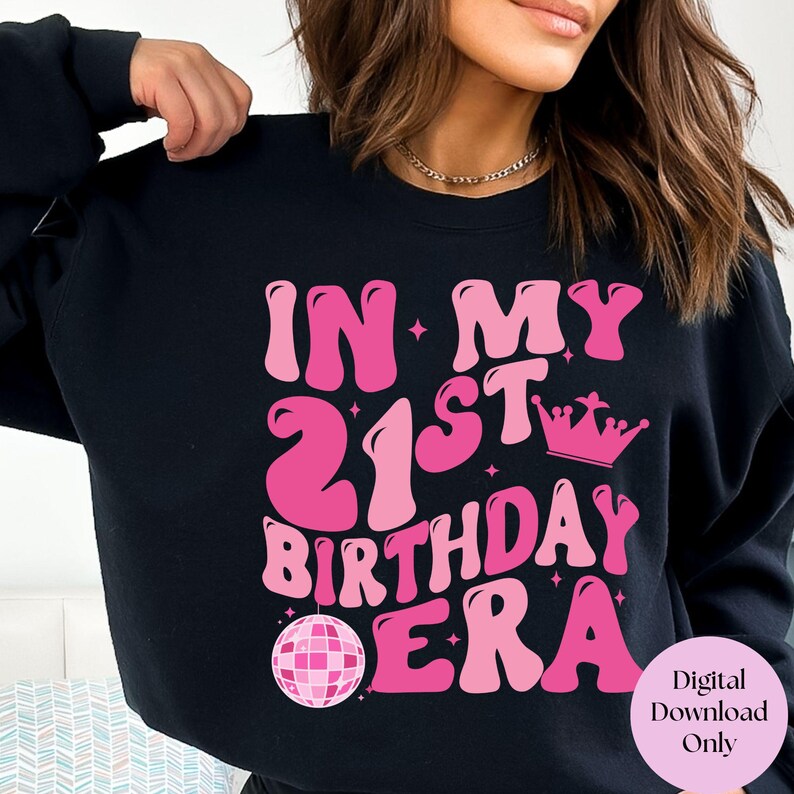 May include: Black sweatshirt with pink text that reads "In My 21st Birthday Era" with a pink disco ball and crown graphic.