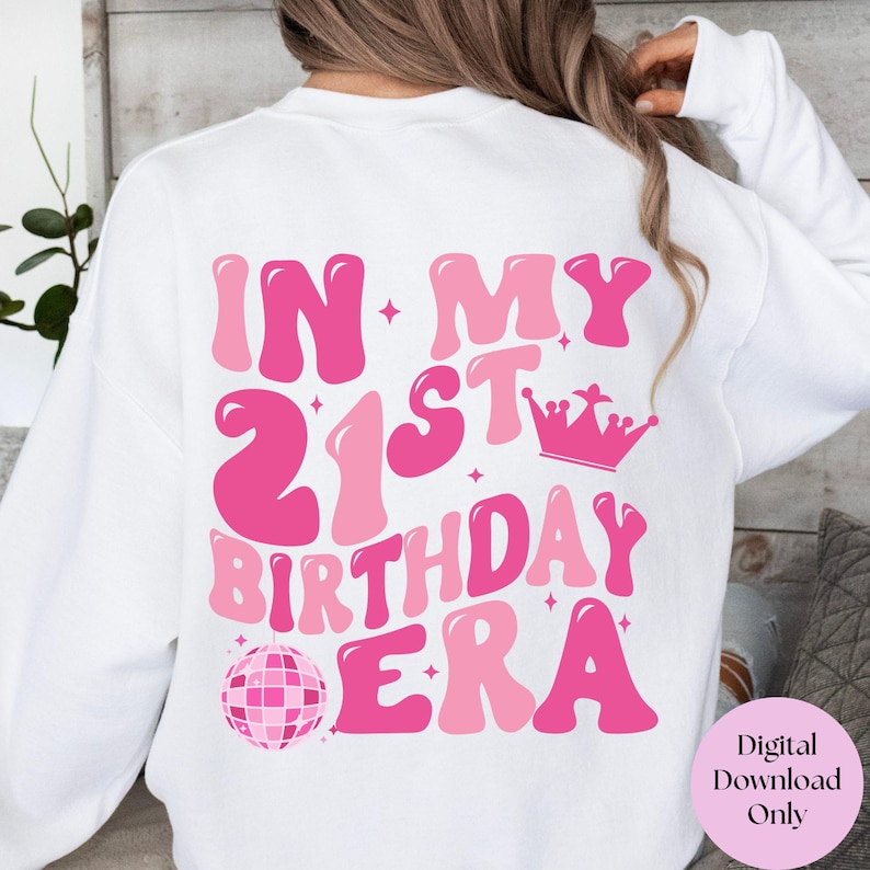 May include: White sweatshirt with pink text that reads "In My 21st Birthday Era" with a pink disco ball and crown graphic.