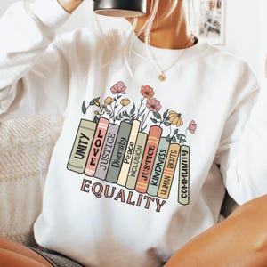 May include: White sweatshirt featuring a graphic of stacked books with words such as "Unity", "Love", "Justice", "Peace", "Inclusion", "Kindness", and "Human Rights". The words are surrounded by flowers and the text "Equality" is printed below the books.