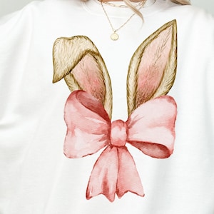 May include: White sweatshirt with a pink bow and brown bunny ears. The bow is tied in a knot and the ears are pointing up.