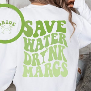 May include: White sweatshirt with green text that reads "Save Water Drink Margs" in a retro font. The text is in a gradient of green shades. A small lime wedge and margarita glass are in the text. A smaller circle on the sweatshirt shows the word "Bride" with two margarita glasses.